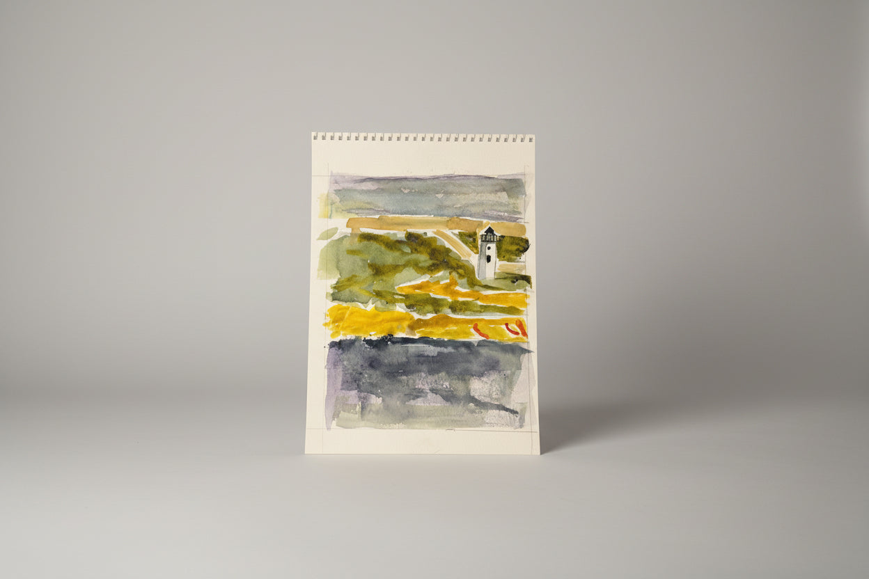 A framed mixed media artwork featuring a lighthouse on paper, displayed on a table with a blurred background of a window and plants.