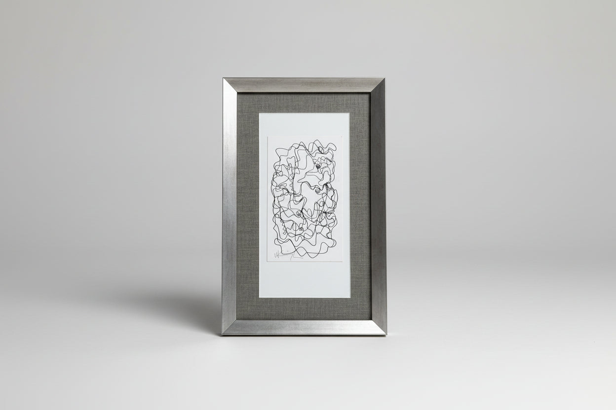 A framed abstract artwork featuring a scribble face pattern in white on a dark background, placed on a wooden surface with a plant and a vase in the background.