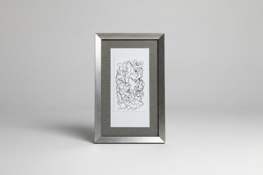 A framed abstract artwork featuring a scribble face pattern in white on a dark background, placed on a wooden surface with a plant and a vase in the background.