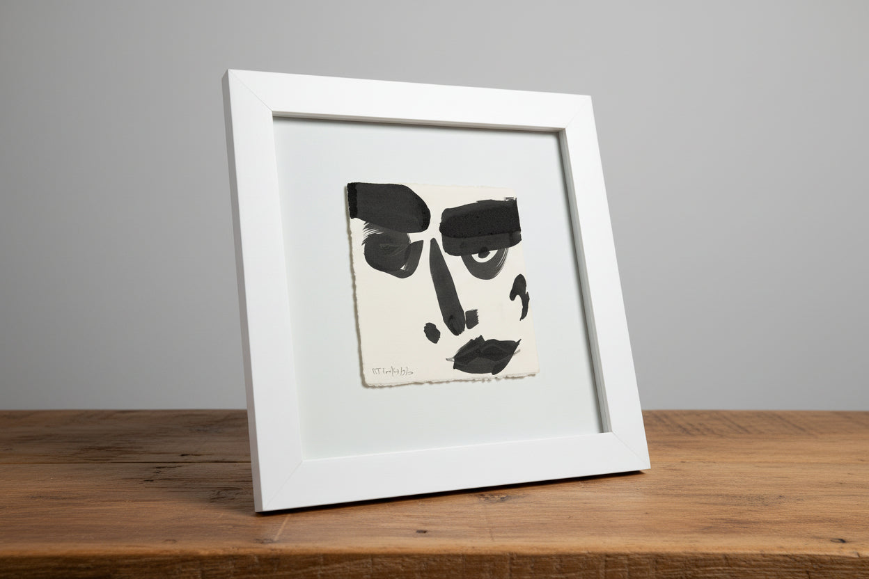 A framed ink portrait of a face displayed on a wooden surface next to a black vase with a flower, with the face painting in the center.