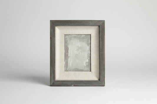 An acrylic on linen panel artwork with a gray wood frame, displayed on a concrete surface with green plants in the background.