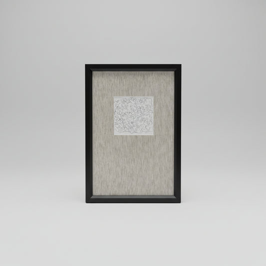 A framed artwork featuring a white background with black scribbles, displayed on a wooden surface with a black vase and a white curtain in the background.