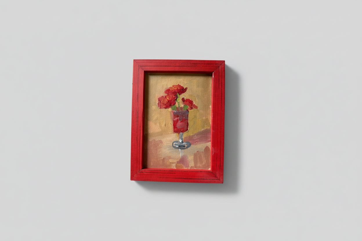 A painting of red flowers in a vase, framed in a red frame, displayed against a wall with plant life around it.