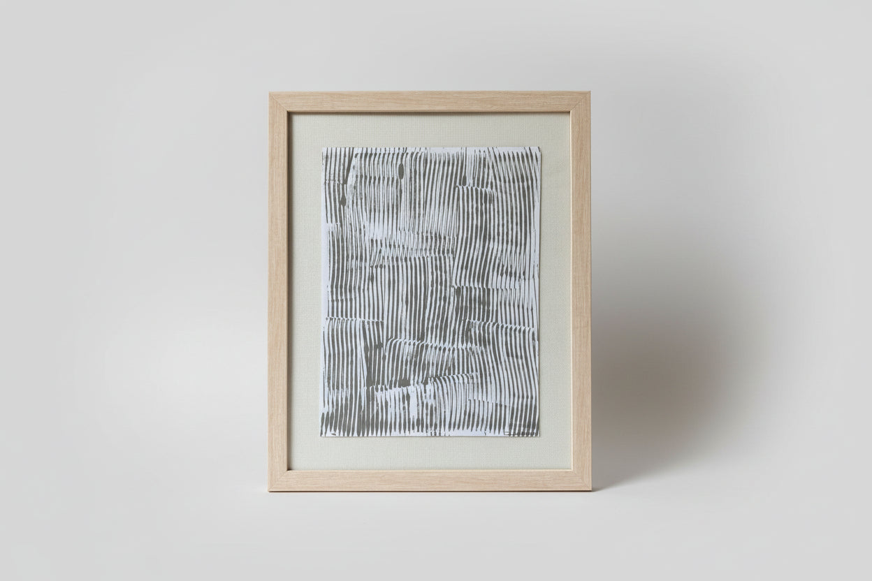 An artwork with abstract figures in a black and white color scheme, framed in a light wood frame, placed on a windowsill with natural lighting in the background.